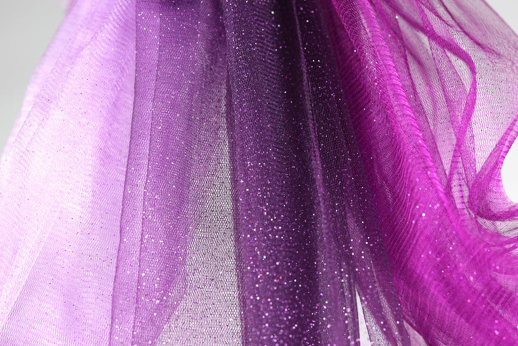 a close-up view of a fabric with a purple and pink color scheme, featuring a glittery texture. The fabric appears to be a sheer or sheer-like material, with a mix of purple and pink hues. The image does not contain any text or other discernible objects or actions.