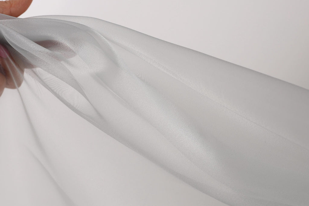 A hand is holding a piece of white fabric, which appears to be a sheer material. The fabric is being held up against a plain white background, creating a simple and minimalist visual composition.