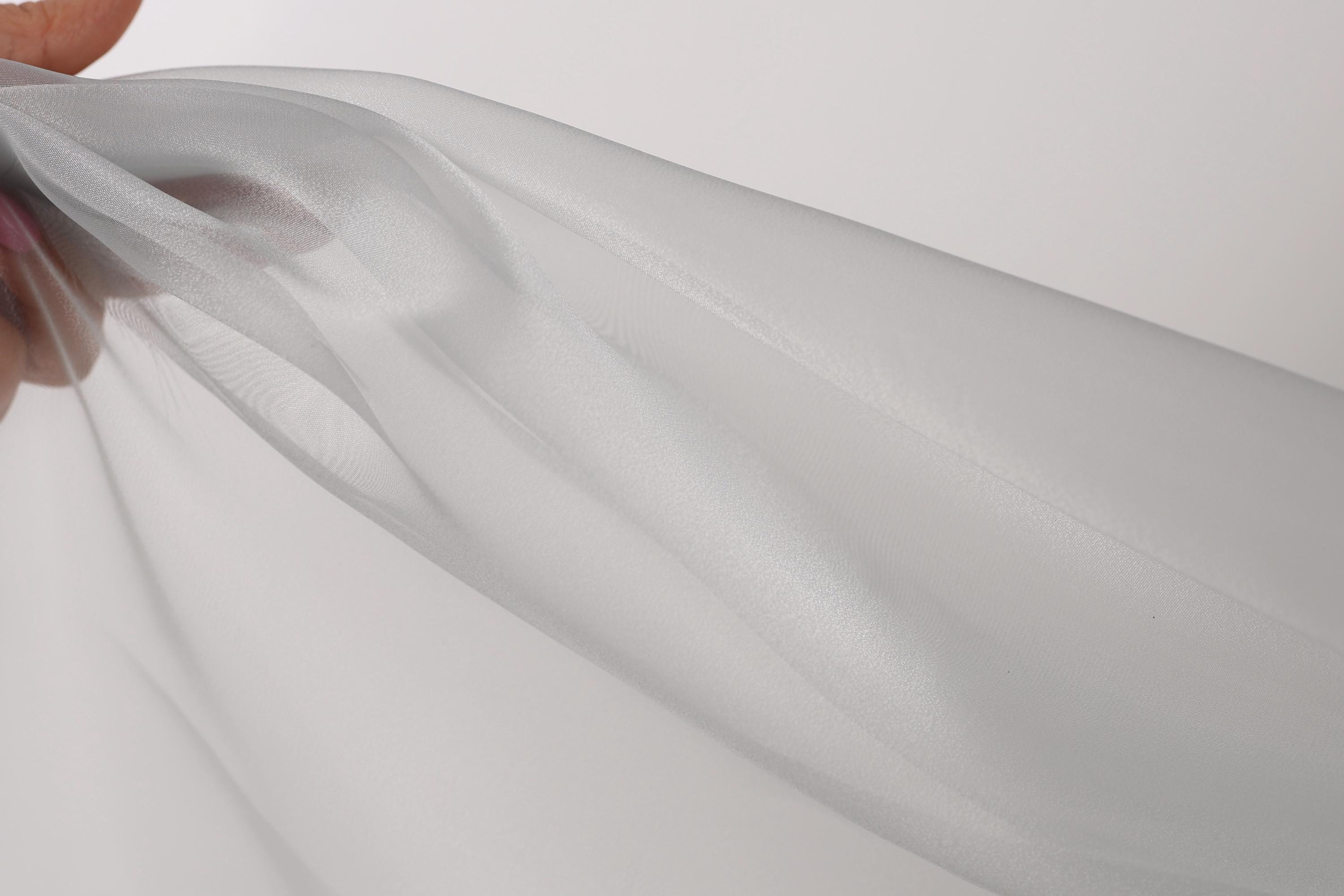 A hand is holding a piece of white fabric, which appears to be a sheer material. The fabric is being held up against a plain white background, creating a simple and minimalist visual composition.