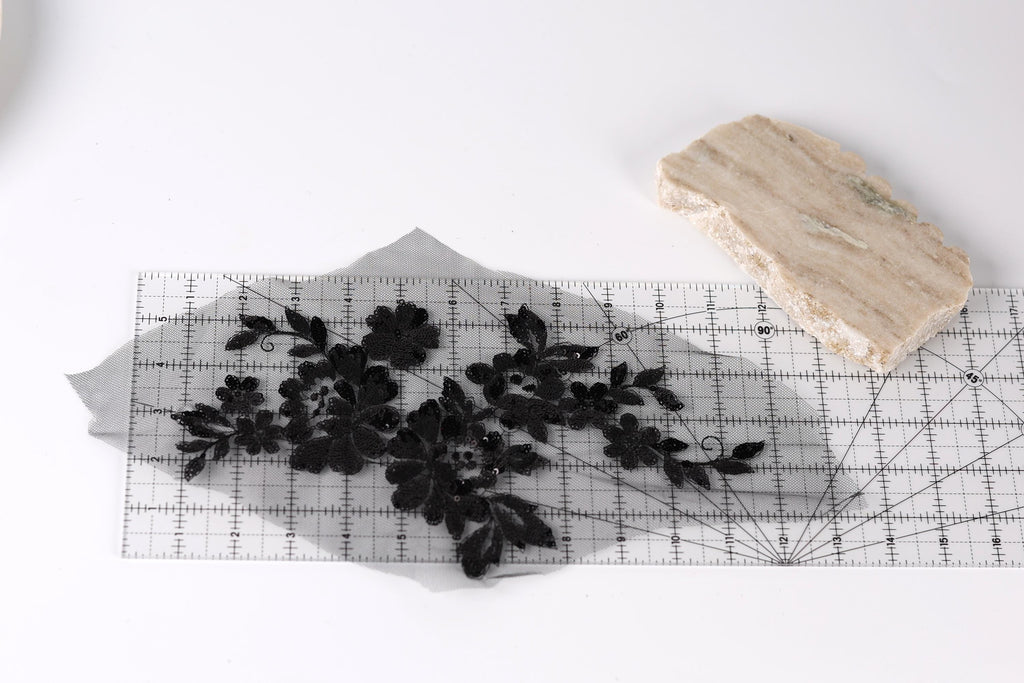 a piece of fabric with black embroidered flowers and leaves, placed on a transparent ruler, which is lying on a white surface.