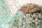 A hand is holding a piece of fabric with intricate floral embroidery, including various shades of green, pink, and white flowers and leaves.