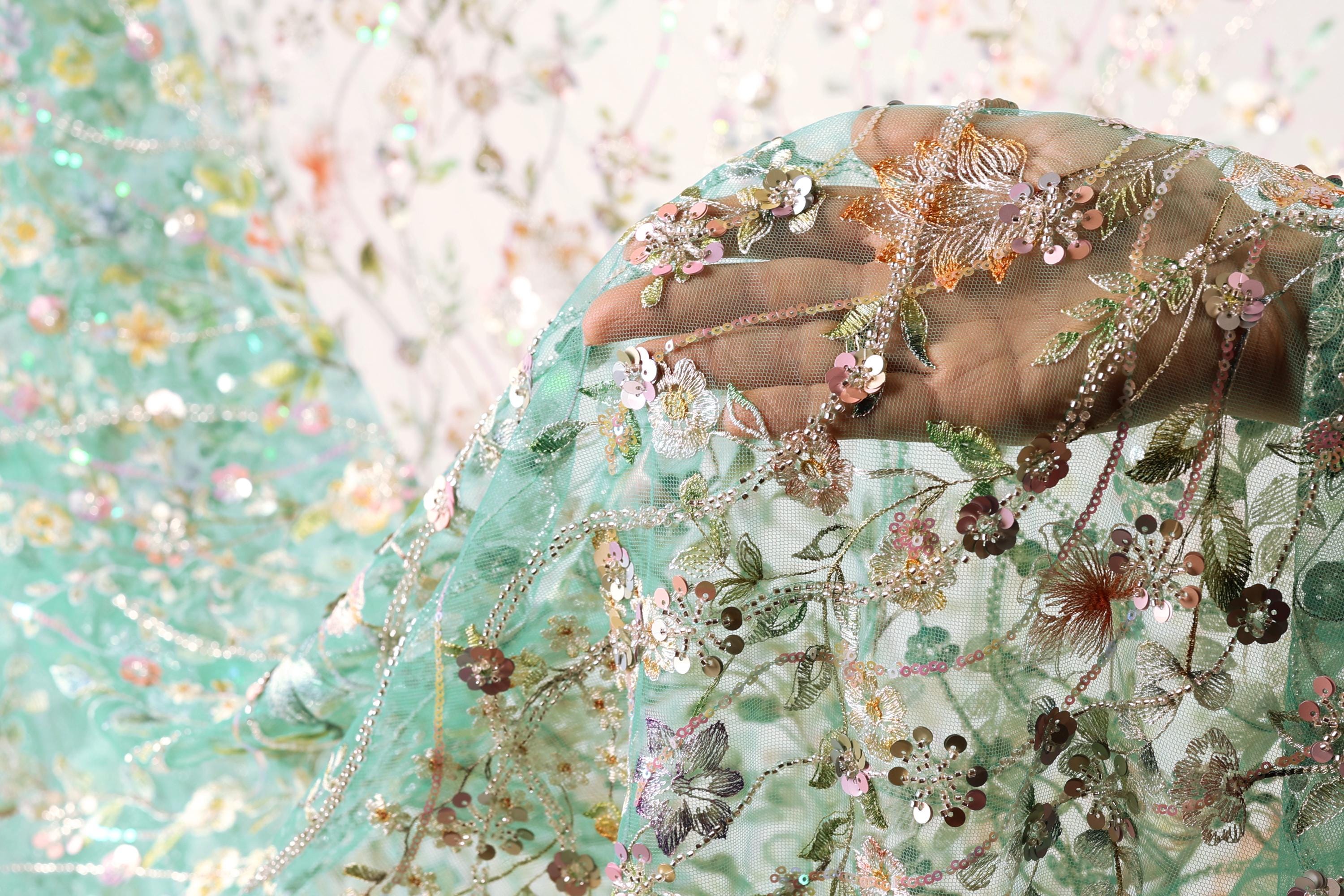 A hand is holding a piece of fabric with intricate floral embroidery, including various shades of green, pink, and white flowers and leaves.