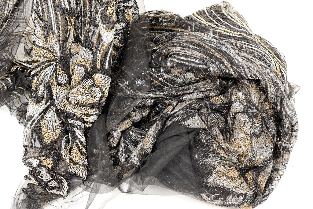 a close-up view of a fabric with a black and gold pattern, featuring a mix of sequins and lace-like elements. The fabric appears to be a sheer material, with a black and gold color scheme.