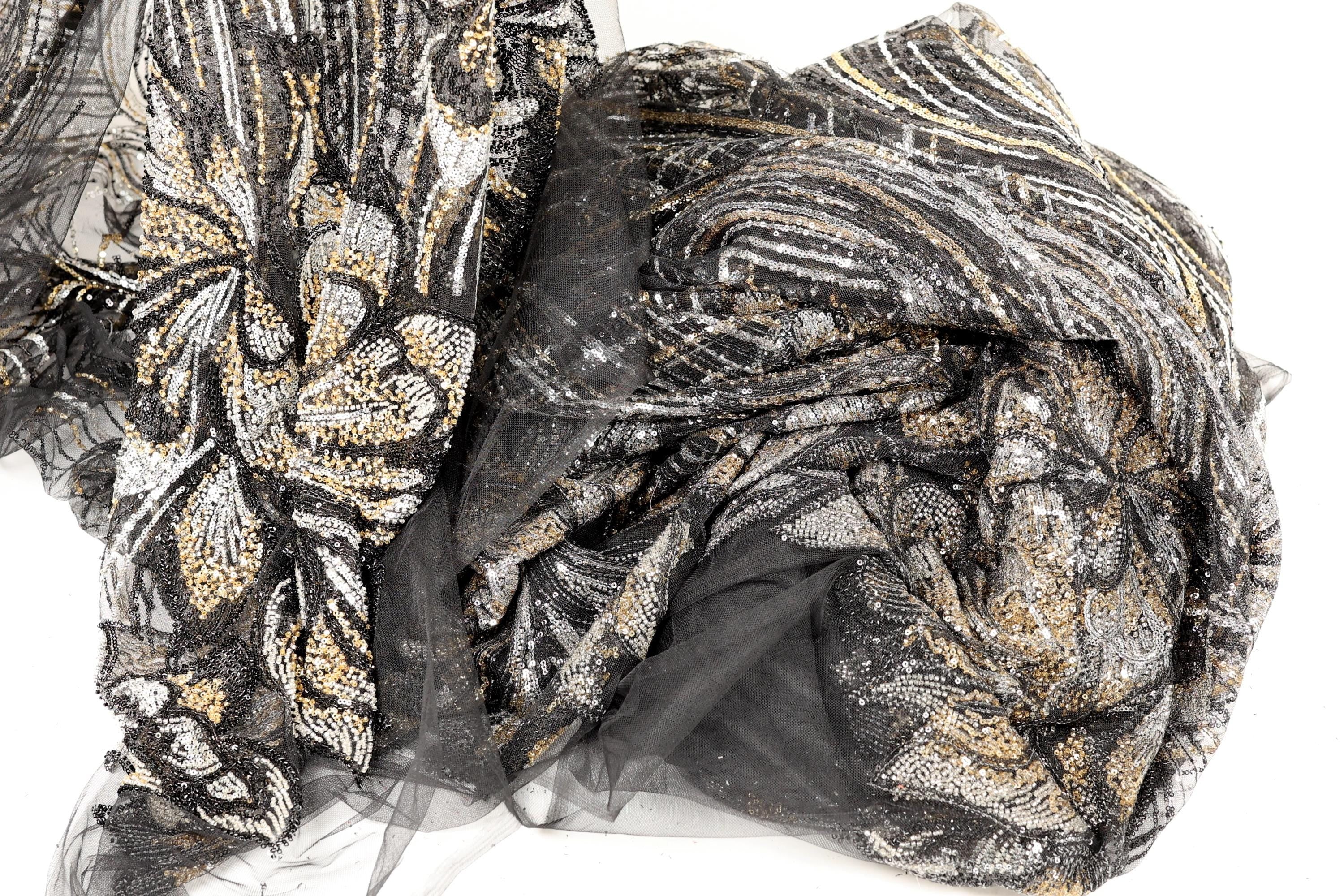 a close-up view of a fabric with a black and gold pattern, featuring a mix of sequins and lace-like elements. The fabric appears to be a sheer material, with a black and gold color scheme.