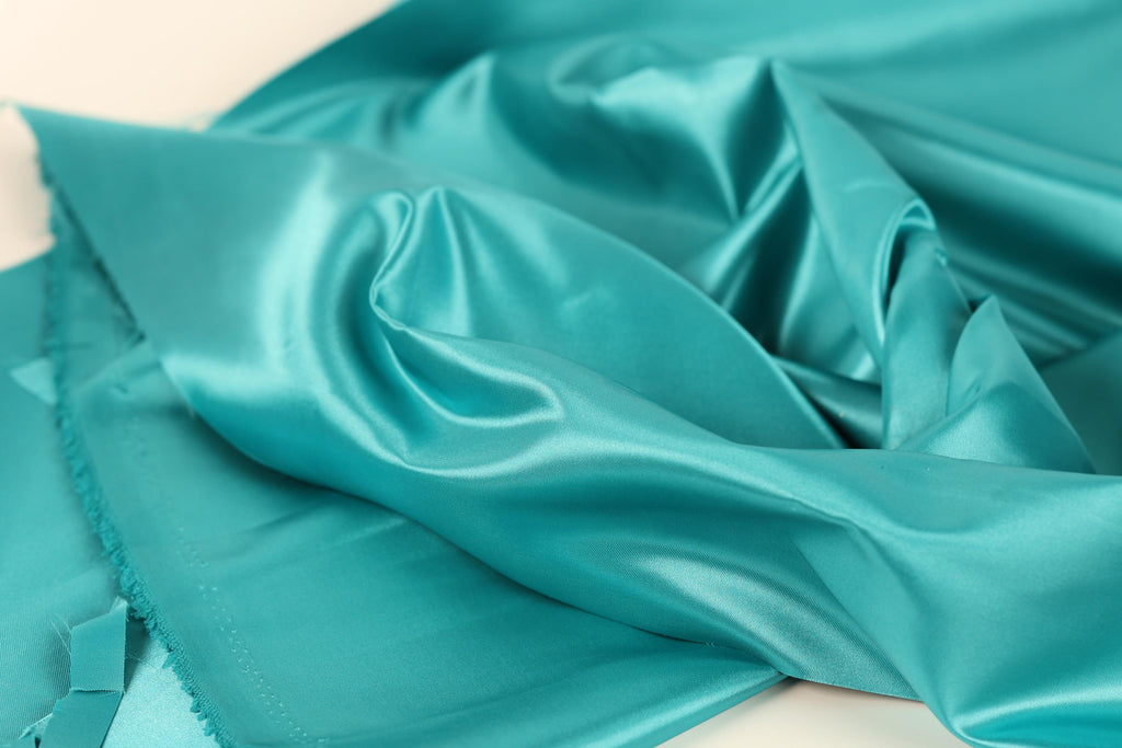 A close-up view of a turquoise satin fabric, with its smooth and shiny texture visible.