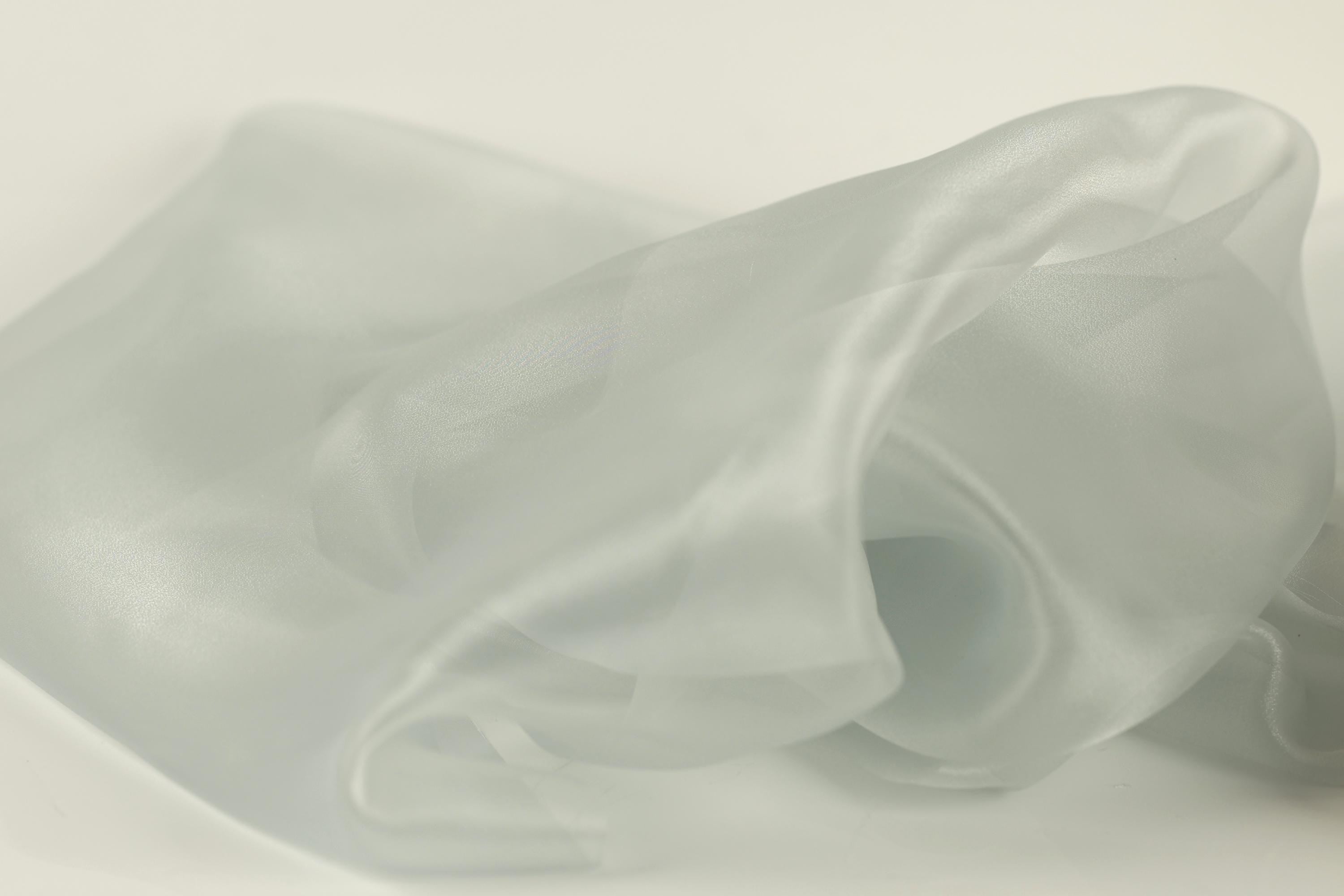 The image is a close-up of a soft, flowing fabric, possibly silk or chiffon, with a light gray or pale green color. The fabric appears to be wrinkled and has a smooth texture, giving it a delicate and elegant appearance.