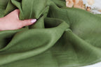 A hand is holding a piece of green fabric, which appears to be made of linen. The fabric is folded and crumpled, giving it a soft and relaxed appearance.
