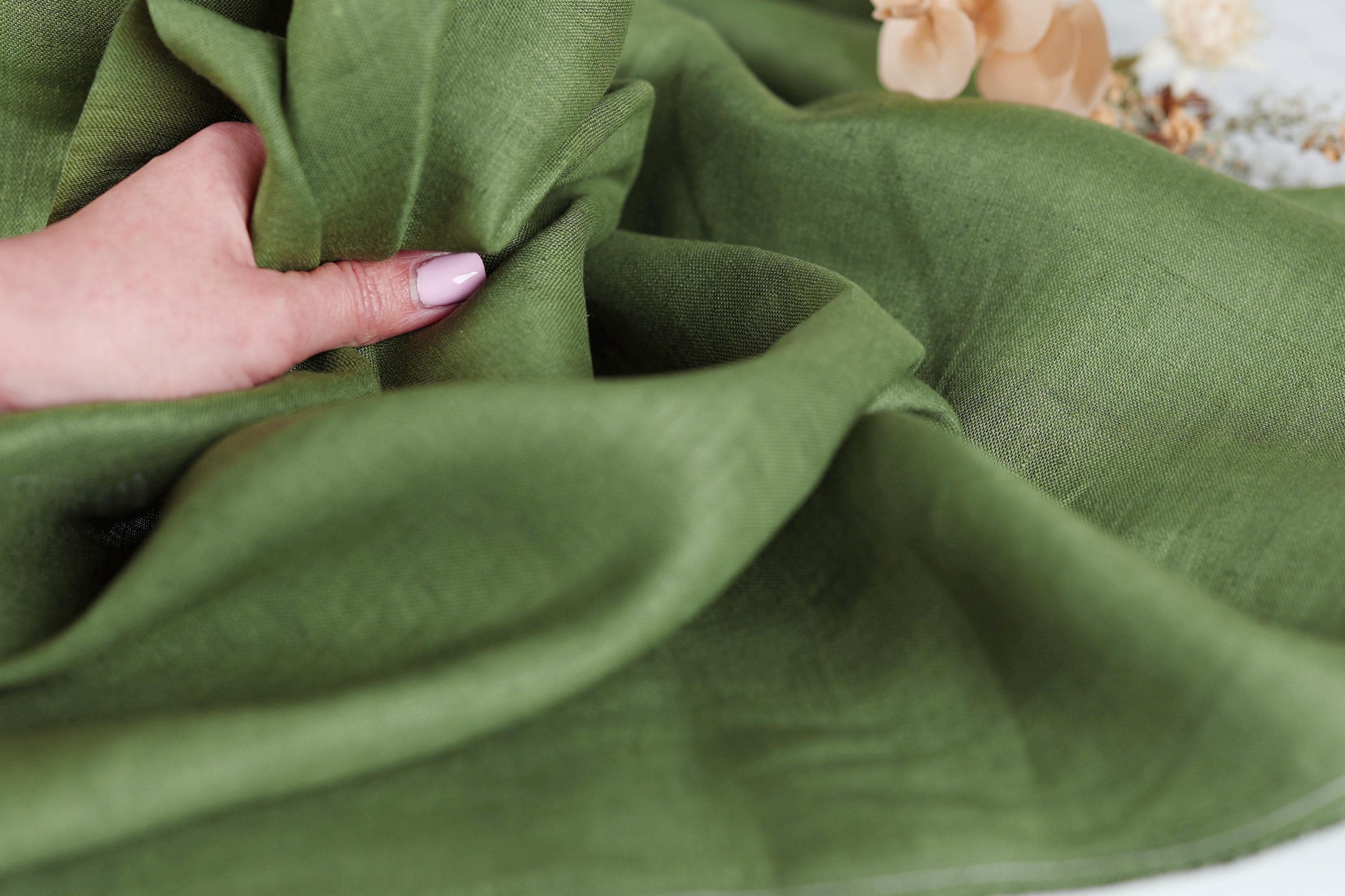 A hand is holding a piece of green fabric, which appears to be made of linen. The fabric is folded and crumpled, giving it a soft and relaxed appearance.