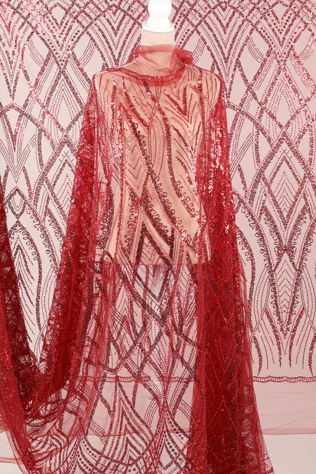 a mannequin wearing a red sequin dress with a sheer overlay. The dress has a long, flowing cape-like design with a red and white pattern. The background is a pink and white patterned fabric.