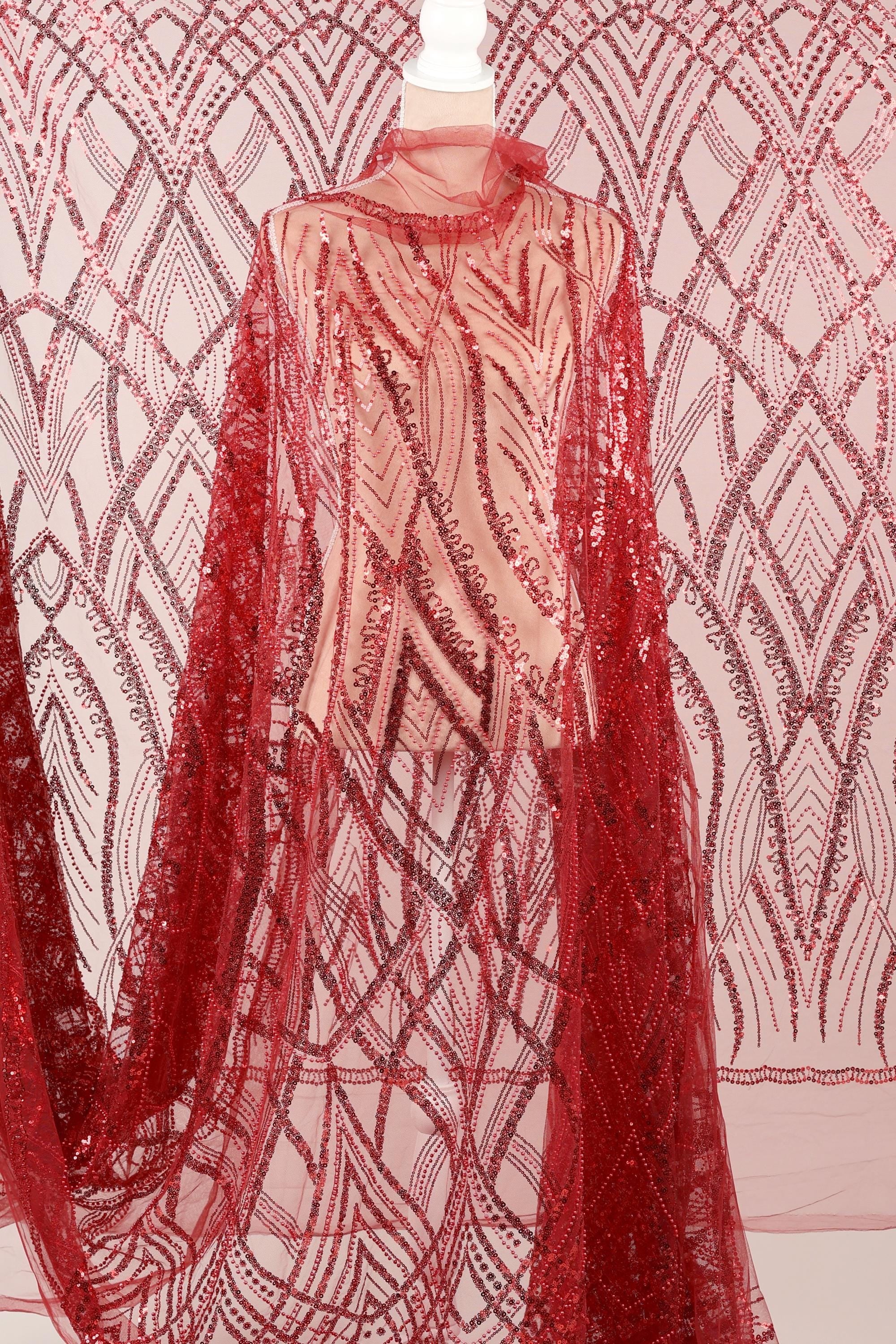 a mannequin wearing a red sequin dress with a sheer overlay. The dress has a long, flowing cape-like design with a red and white pattern. The background is a pink and white patterned fabric.