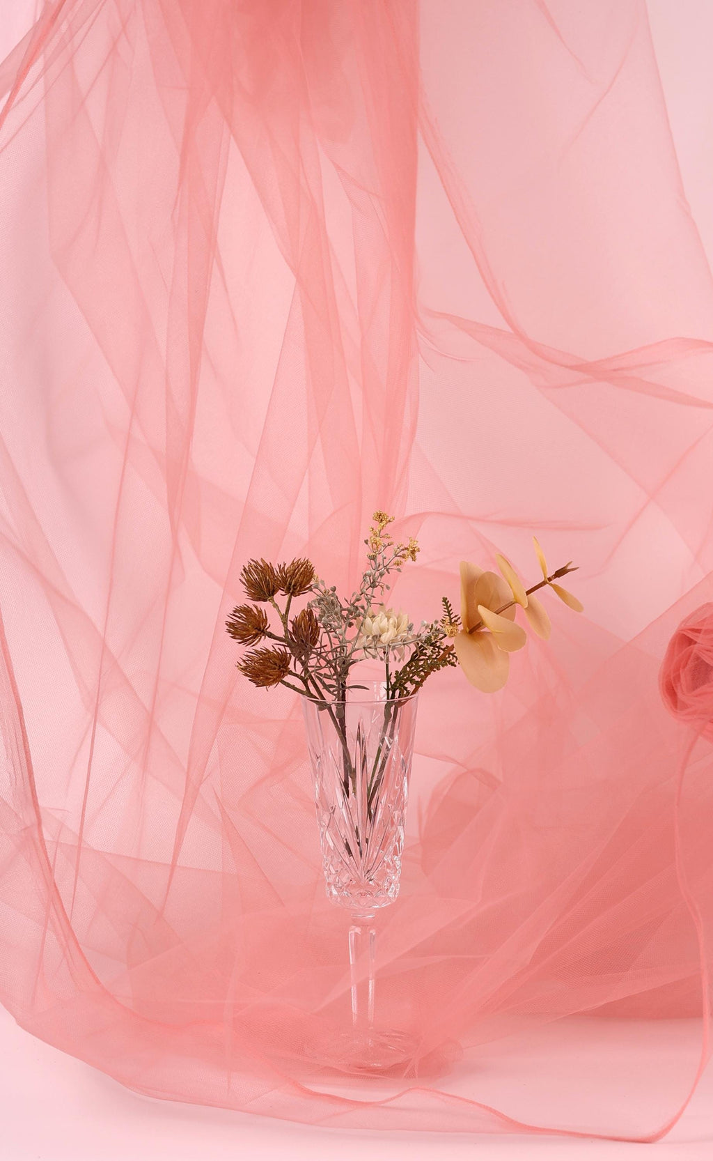 A clear glass vase filled with dried flowers and branches sits on a pink background, surrounded by a flowing pink fabric.