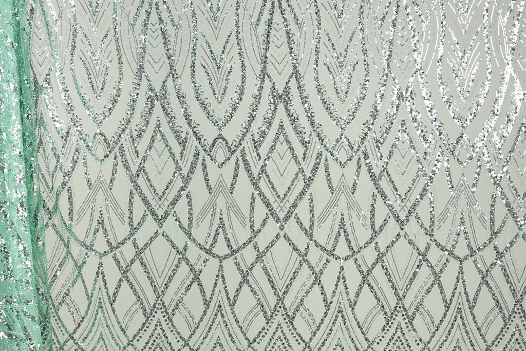 a close-up view of a fabric with a repeating pattern of silver and green sequin designs. The pattern consists of a series of diamond shapes and lines, creating a visually striking and intricate design. The fabric appears to be a light green color, which complements the silver and green sequins. The image does not contain any text or other discernible objects, and the focus is solely on the detailed pattern of the sequin design.