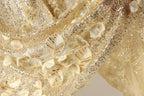 The image is a close-up of a fabric with gold sequins and delicate white flowers, creating a shimmering and delicate appearance.