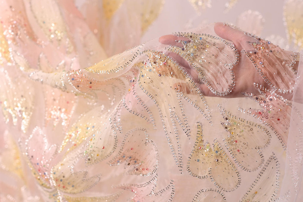 A close-up view of a delicate, hand-embroidered fabric with a floral pattern. The fabric appears to be made of sheer material, with a light pink and white color scheme. The intricate embroidery and beading on the fabric create a visually appealing and delicate appearance.