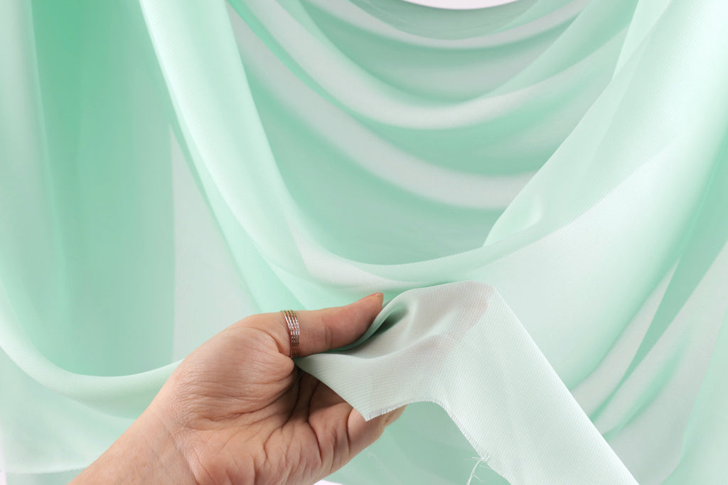 A hand is holding a piece of light green fabric, possibly silk, against a background of a similar light green fabric.