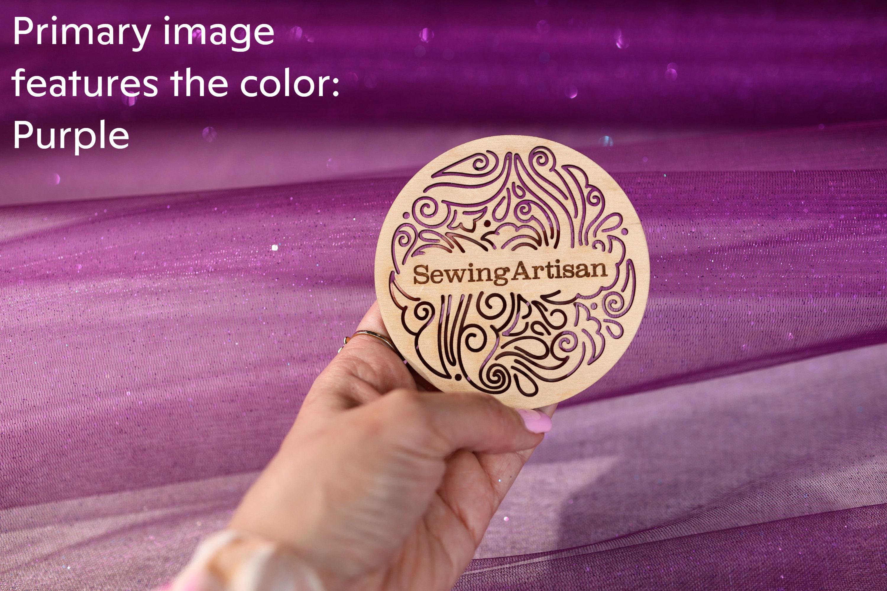 A hand holding a wooden coaster with the text "SewingArtisan" on it, against a purple background.