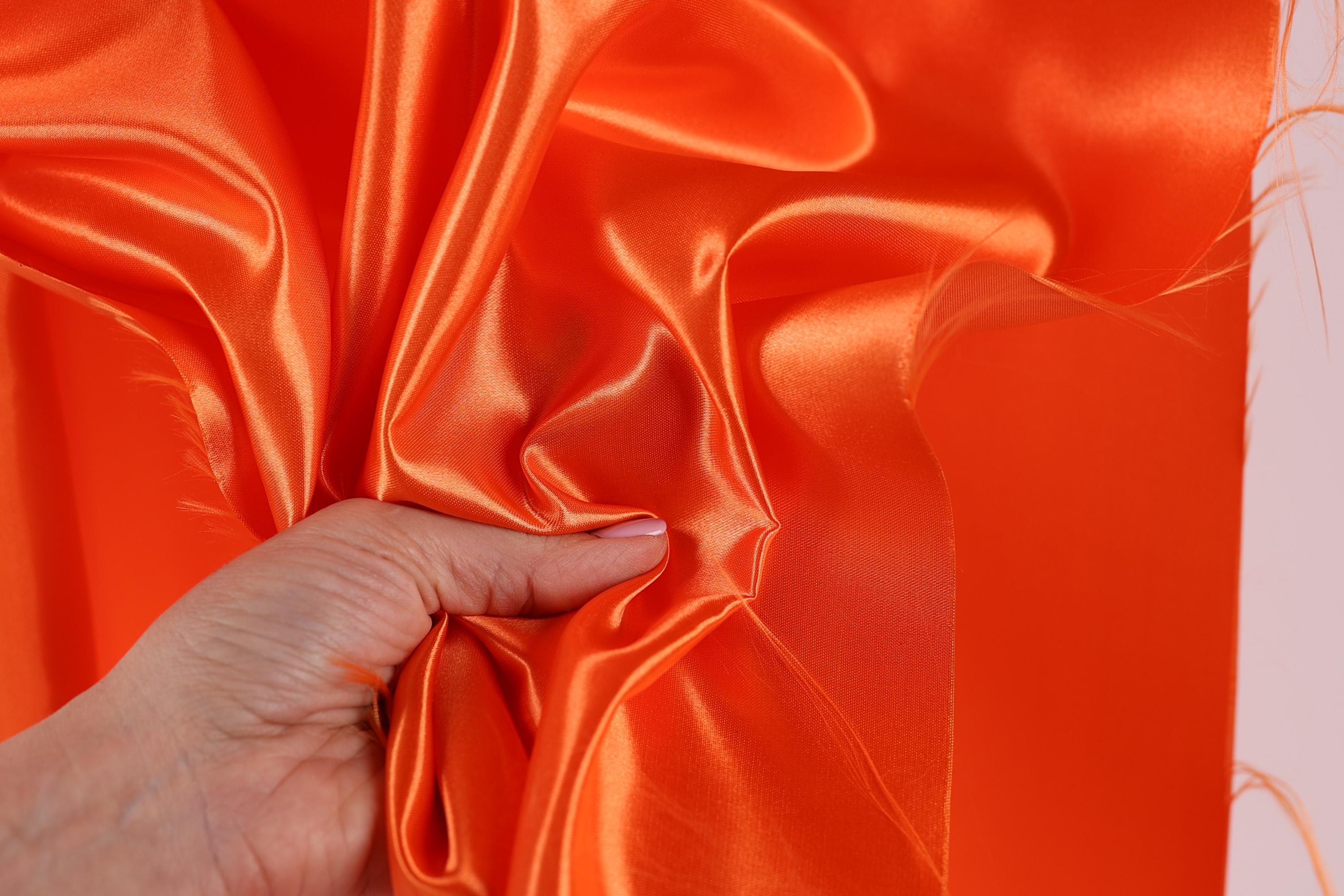 A hand is holding a piece of orange satin fabric, which appears to be soft and shiny.
