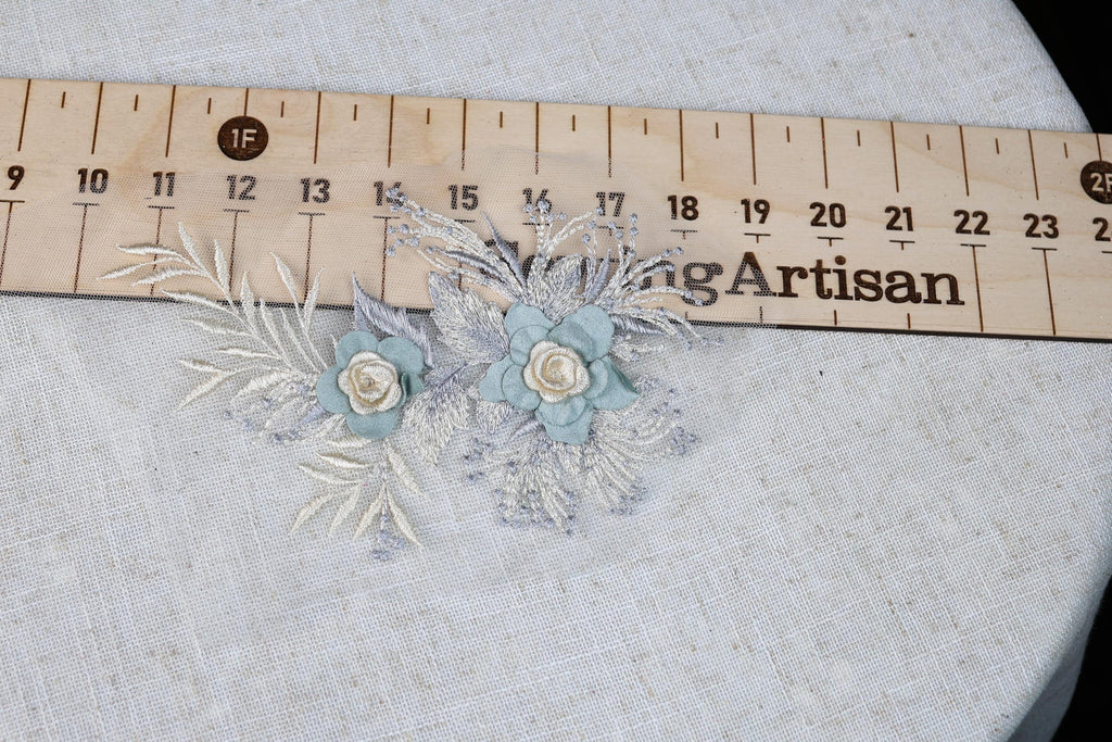 a close-up view of a floral embroidery with two blue flowers and some white leaves, placed on a white fabric surface. A wooden ruler is visible in the background, indicating the size or measurement of the embroidery.