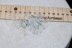 a close-up view of a floral embroidery with two blue flowers and some white leaves, placed on a white fabric surface. A wooden ruler is visible in the background, indicating the size or measurement of the embroidery.