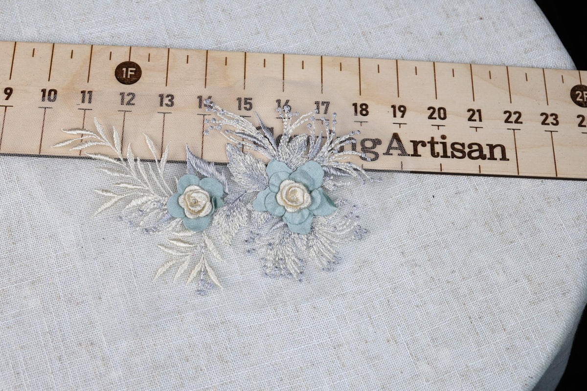 a close-up view of a floral embroidery with two blue flowers and some white leaves, placed on a white fabric surface. A wooden ruler is visible in the background, indicating the size or measurement of the embroidery.
