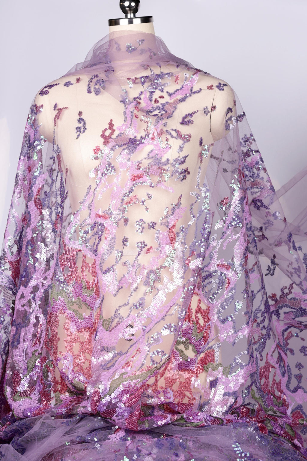 a mannequin wearing a sheer, flowy fabric with a floral pattern in shades of purple and pink. The fabric appears to be made of sequins, giving it a shiny and decorative appearance.