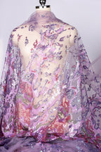 a mannequin wearing a sheer, flowy fabric with a floral pattern in shades of purple and pink. The fabric appears to be made of sequins, giving it a shiny and decorative appearance.
