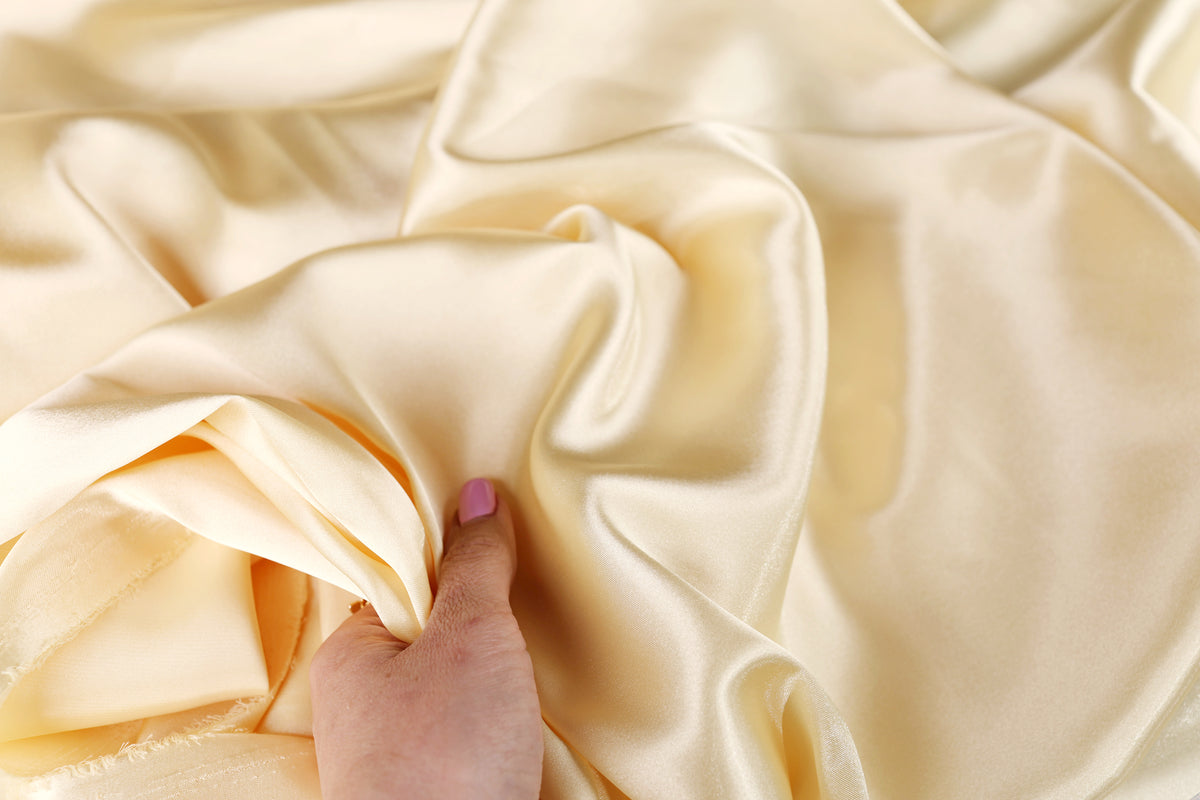 A hand is holding a piece of light yellow satin fabric, which appears to be soft and smooth.