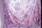 a close-up view of a fabric with a floral pattern in shades of purple and pink. The fabric appears to be made of a sheer material, with a sheer curtain visible in the background.