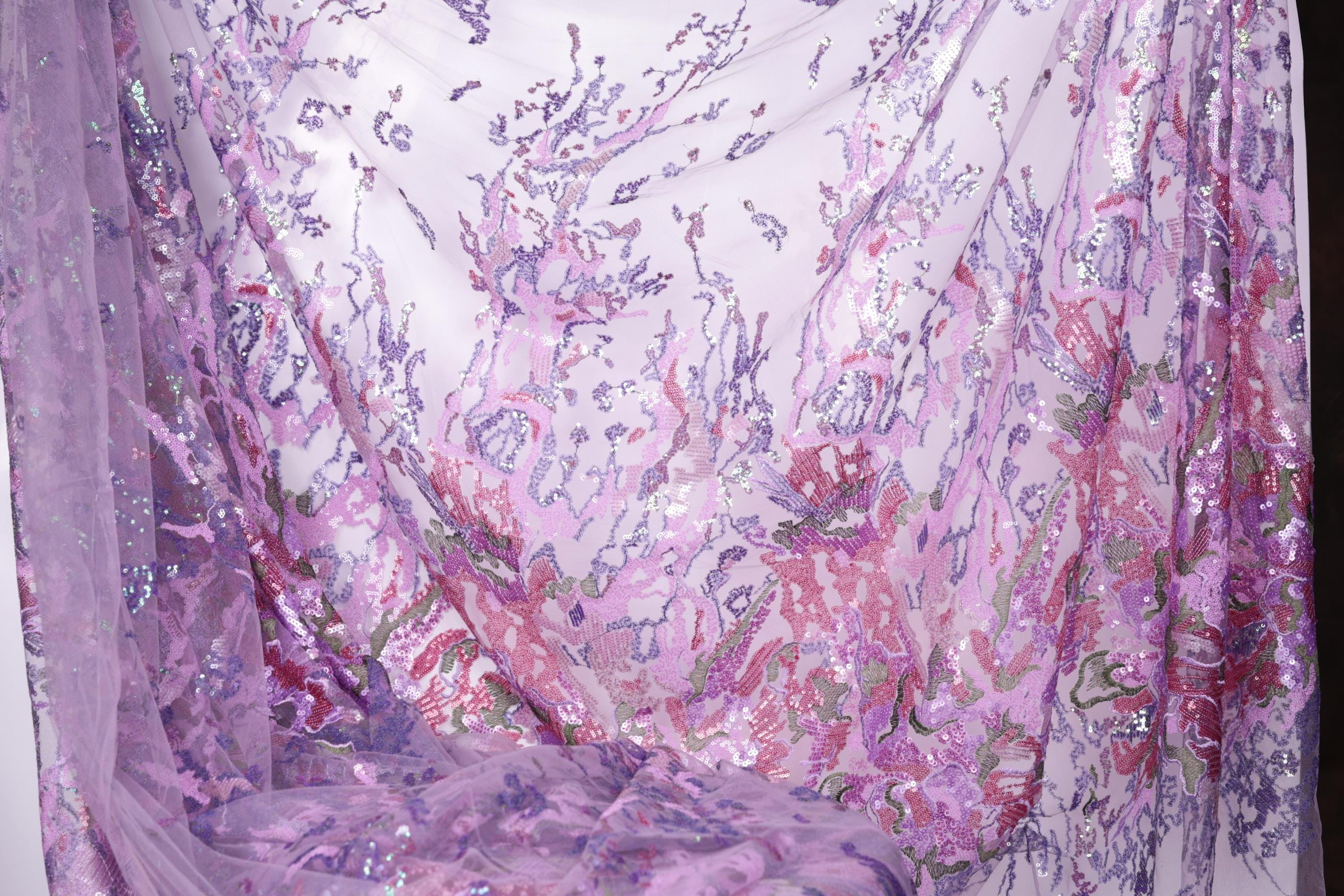 a close-up view of a fabric with a floral pattern in shades of purple and pink. The fabric appears to be made of a sheer material, with a sheer curtain visible in the background.