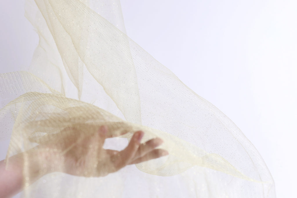 A hand is holding a piece of light-colored fabric, possibly a sheer or sheer-like material, against a plain white background.