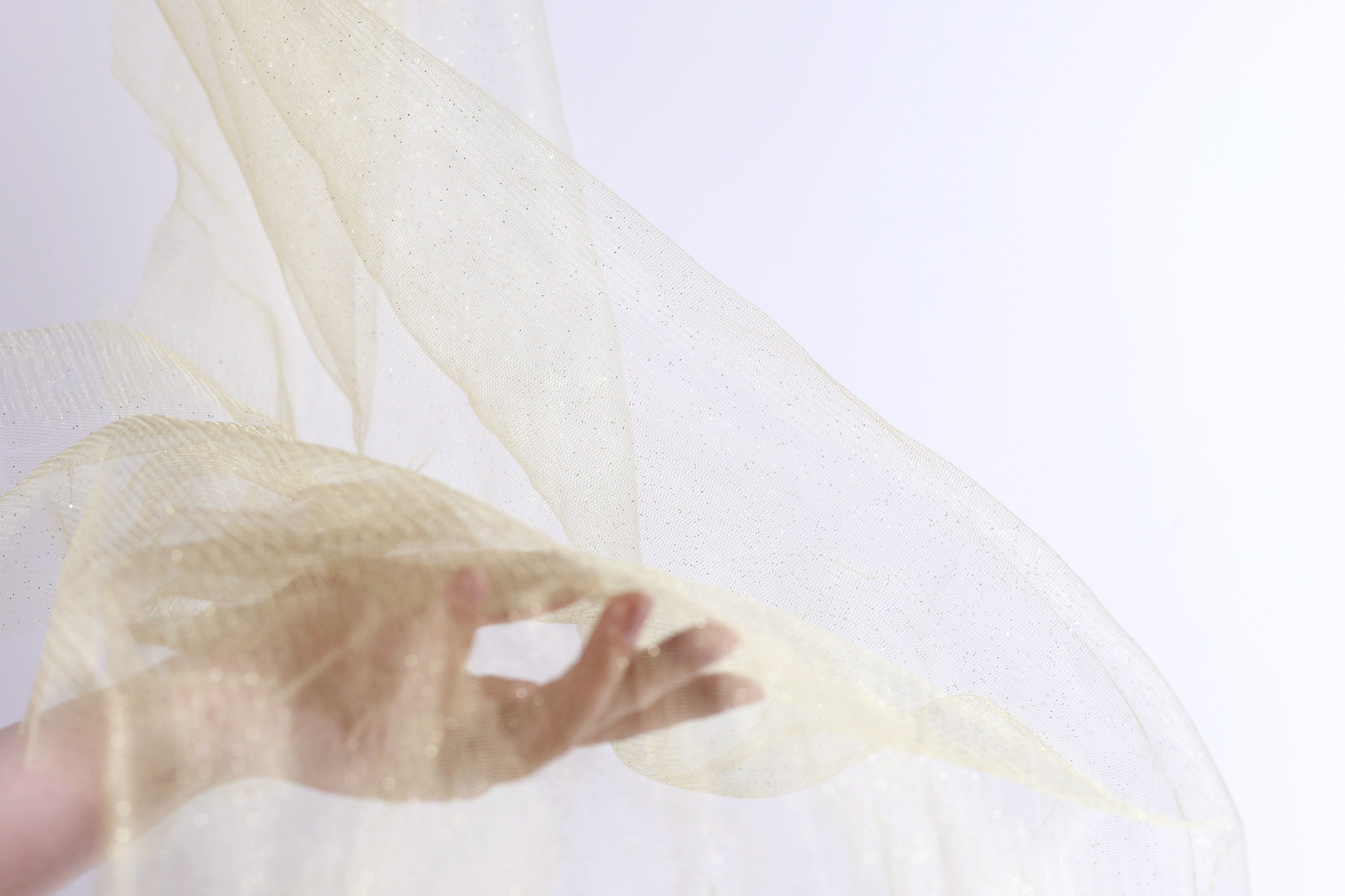 A hand is holding a piece of light-colored fabric, possibly a sheer or sheer-like material, against a plain white background.
