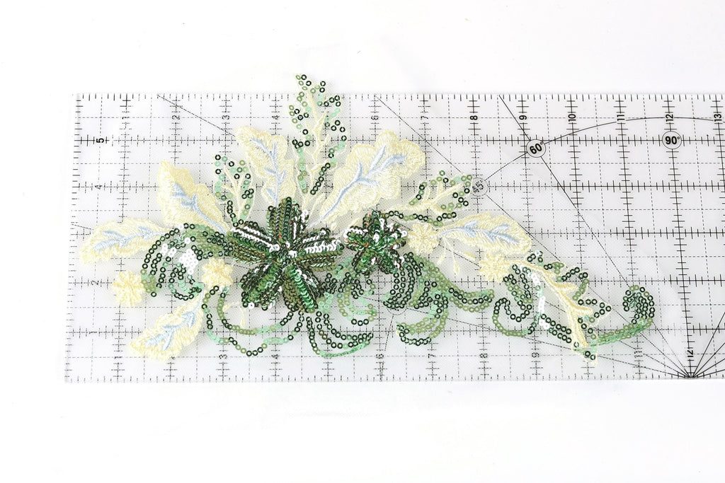 a decorative piece of embroidered fabric with green and white floral elements, placed on a transparent ruler for scale.