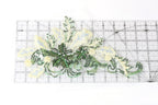 a decorative piece of embroidered fabric with green and white floral elements, placed on a transparent ruler for scale.