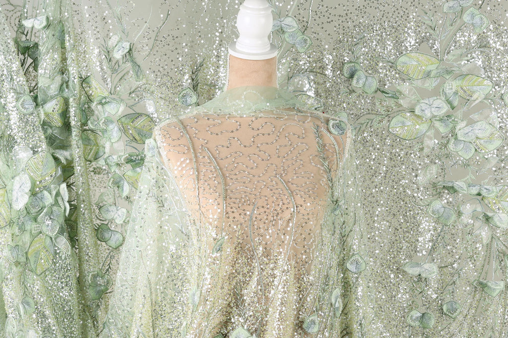 a mannequin wearing a sheer, green and white floral fabric draped over it. The fabric has a shimmering, sequined appearance, creating a visually striking and elegant effect.