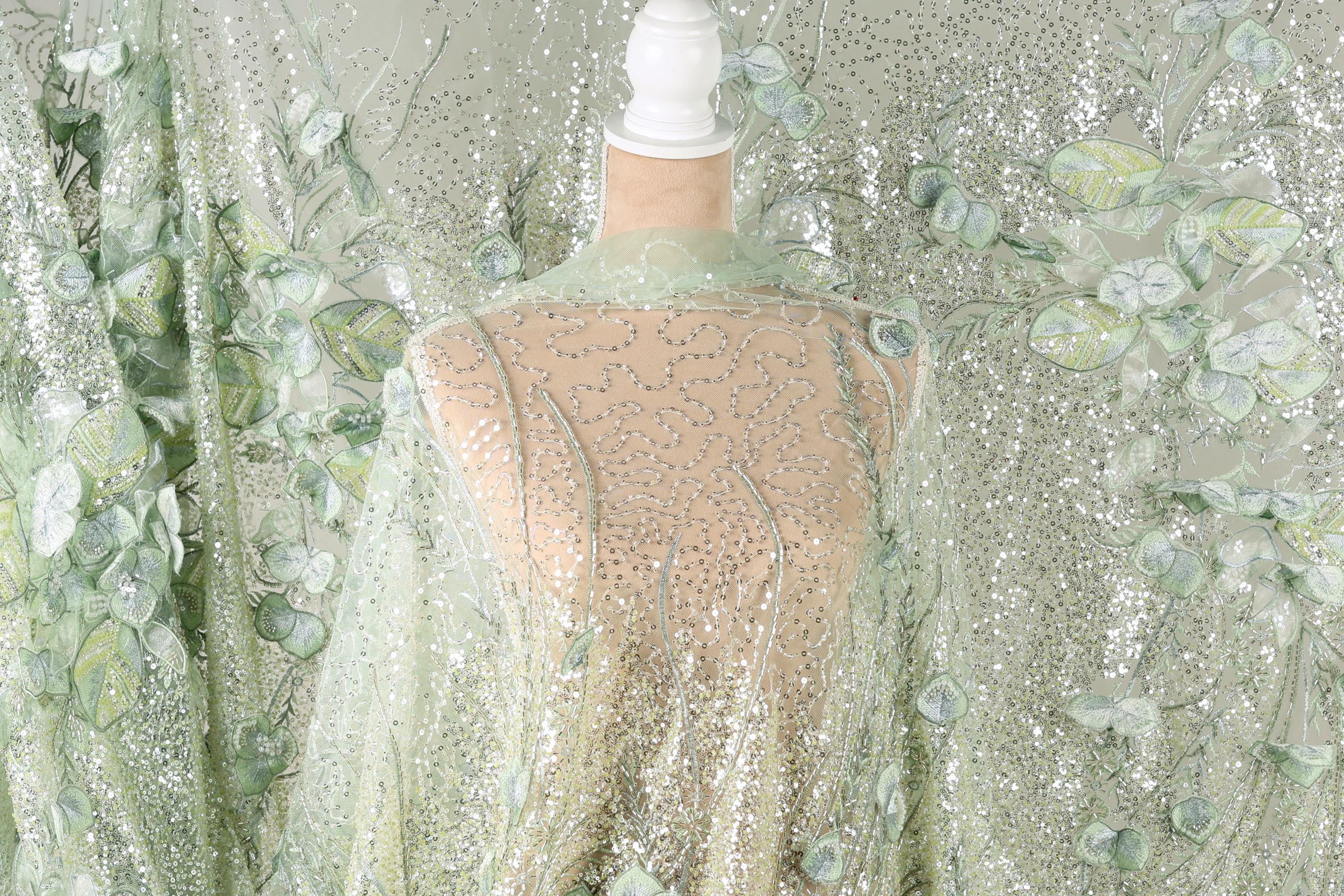 a mannequin wearing a sheer, green and white floral fabric draped over it. The fabric has a shimmering, sequined appearance, creating a visually striking and elegant effect.