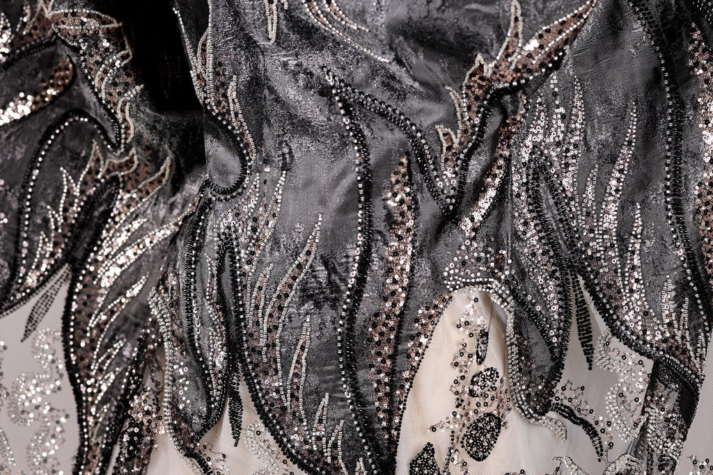 The image is a close-up view of a fabric with intricate black and silver embroidery, featuring a floral pattern and sparkling embellishments.