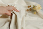 A hand is holding a piece of light-colored fabric, with a blurred background featuring a bouquet of yellow flowers.