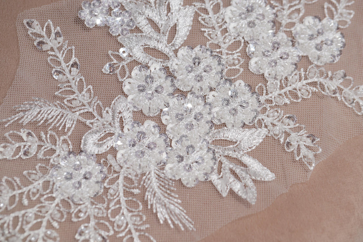 a close-up view of a delicate, intricate white lace fabric with a floral pattern. The lace appears to be made of a fine, sheer material, and the intricate details of the lace design are clearly visible.