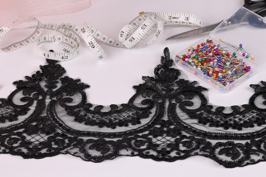 a close-up view of a black lace trim with intricate floral patterns, along with a spool of thread, a pair of scissors, and a container of colorful pins.