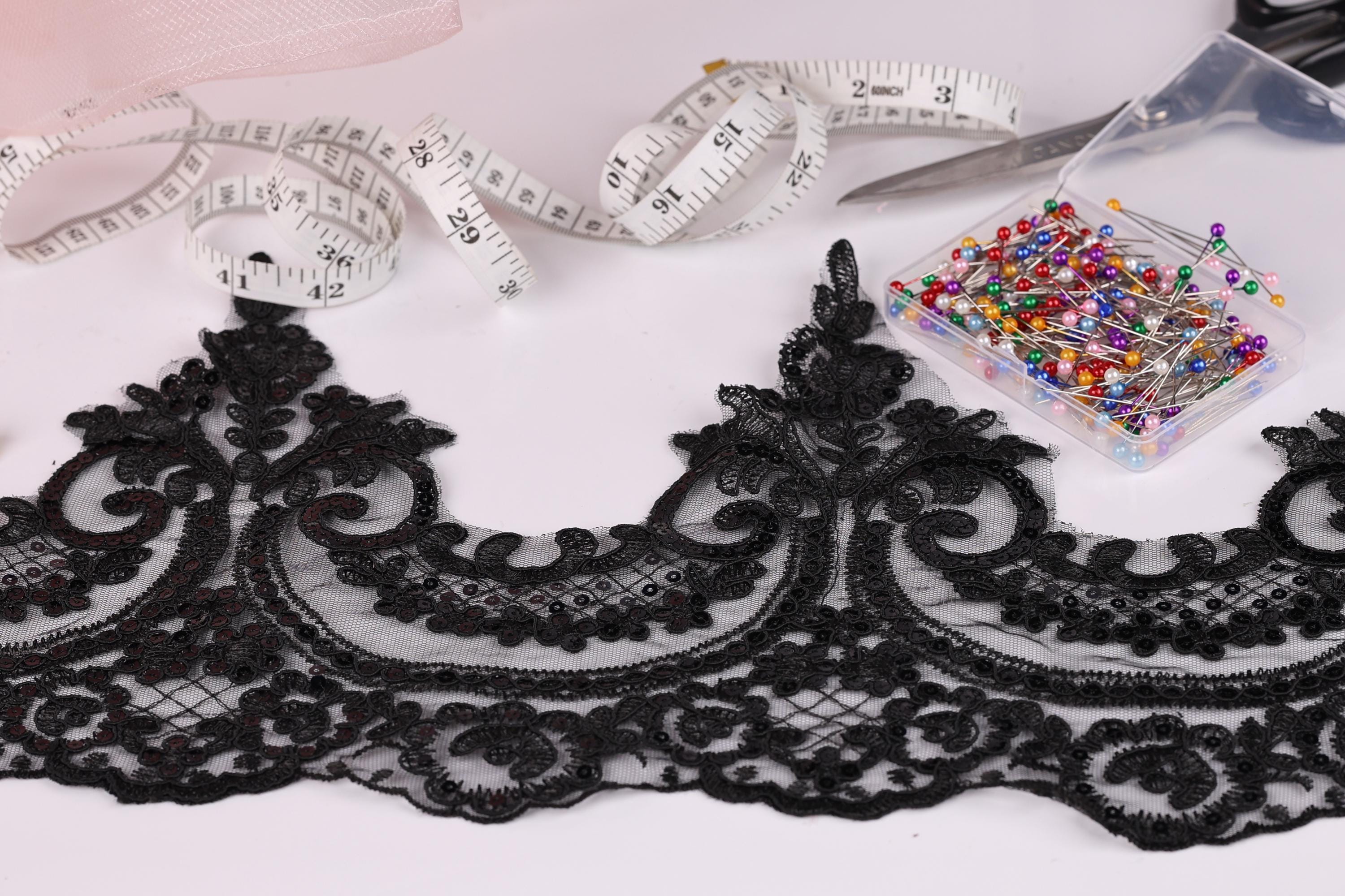 a close-up view of a black lace trim with intricate floral patterns, along with a spool of thread, a pair of scissors, and a container of colorful pins.
