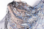 a close-up view of a sheer, sheer fabric with a floral pattern. The fabric appears to be made of sheer material, giving it a delicate and flowing appearance. The floral pattern consists of various shades of blue, gray, and white, creating a visually striking and intricate design. The fabric is spread out, revealing its full and detailed appearance.