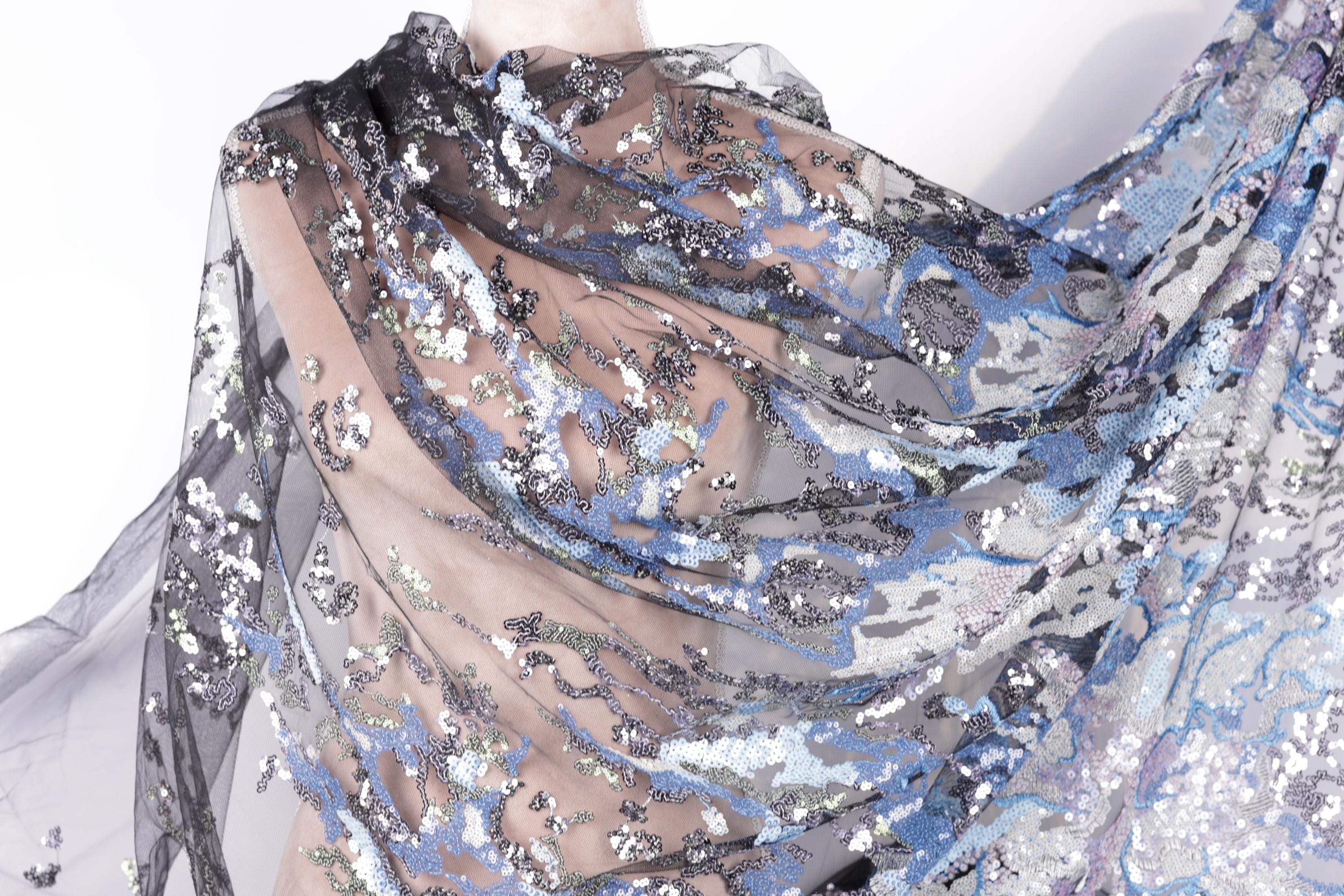 a close-up view of a sheer, sheer fabric with a floral pattern. The fabric appears to be made of sheer material, giving it a delicate and flowing appearance. The floral pattern consists of various shades of blue, gray, and white, creating a visually striking and intricate design. The fabric is spread out, revealing its full and detailed appearance.