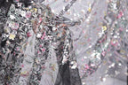 The image is a close-up view of a delicate, intricately embroidered fabric with a floral pattern. The fabric appears to be a sheer material, with a black background that contrasts with the colorful floral design. The image is not a collage, but a single, focused shot of the fabric, showcasing its detailed embroidery and the intricate patterns created by the flowers and other embellishments.