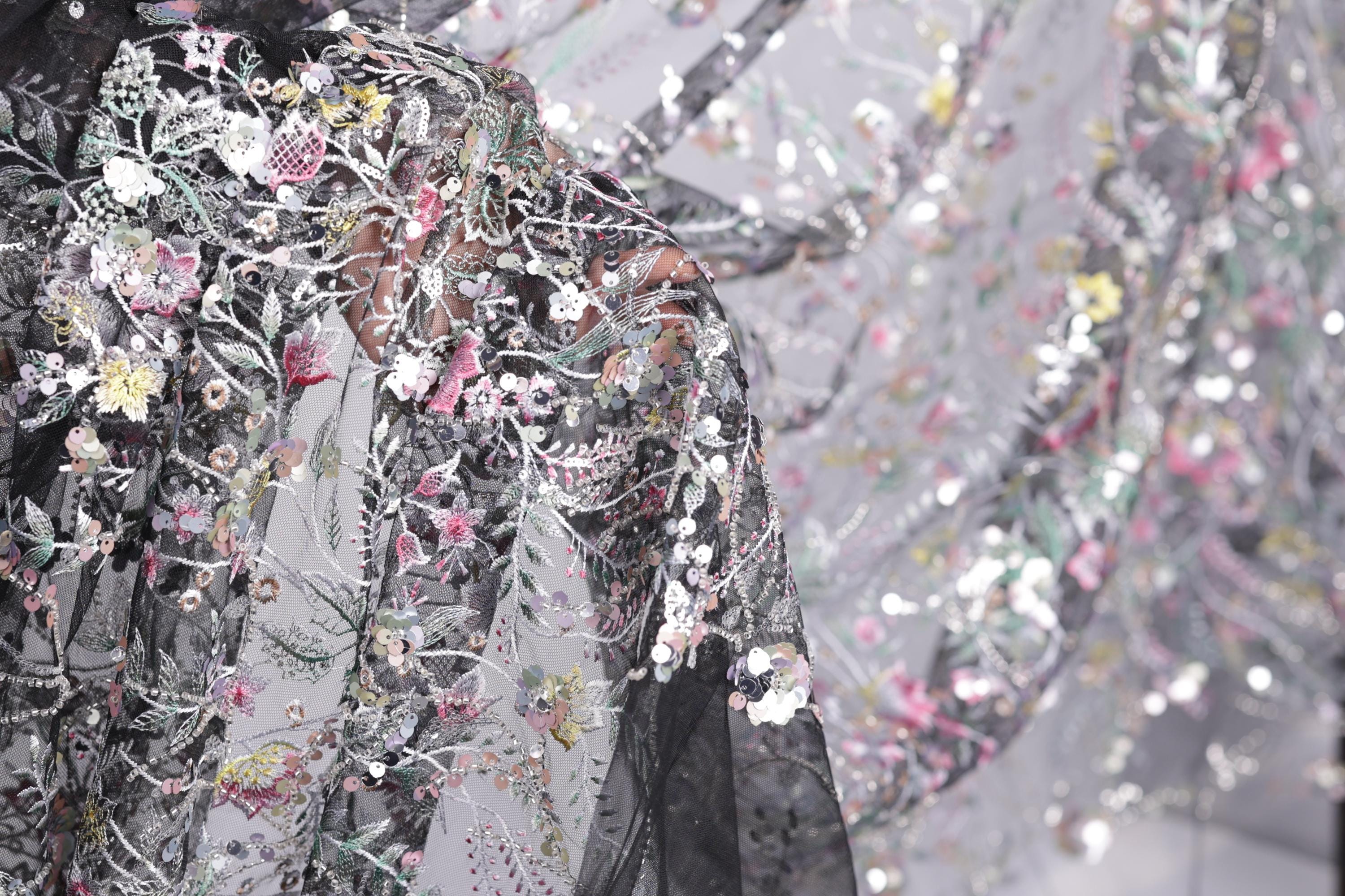 The image is a close-up view of a delicate, intricately embroidered fabric with a floral pattern. The fabric appears to be a sheer material, with a black background that contrasts with the colorful floral design. The image is not a collage, but a single, focused shot of the fabric, showcasing its detailed embroidery and the intricate patterns created by the flowers and other embellishments.