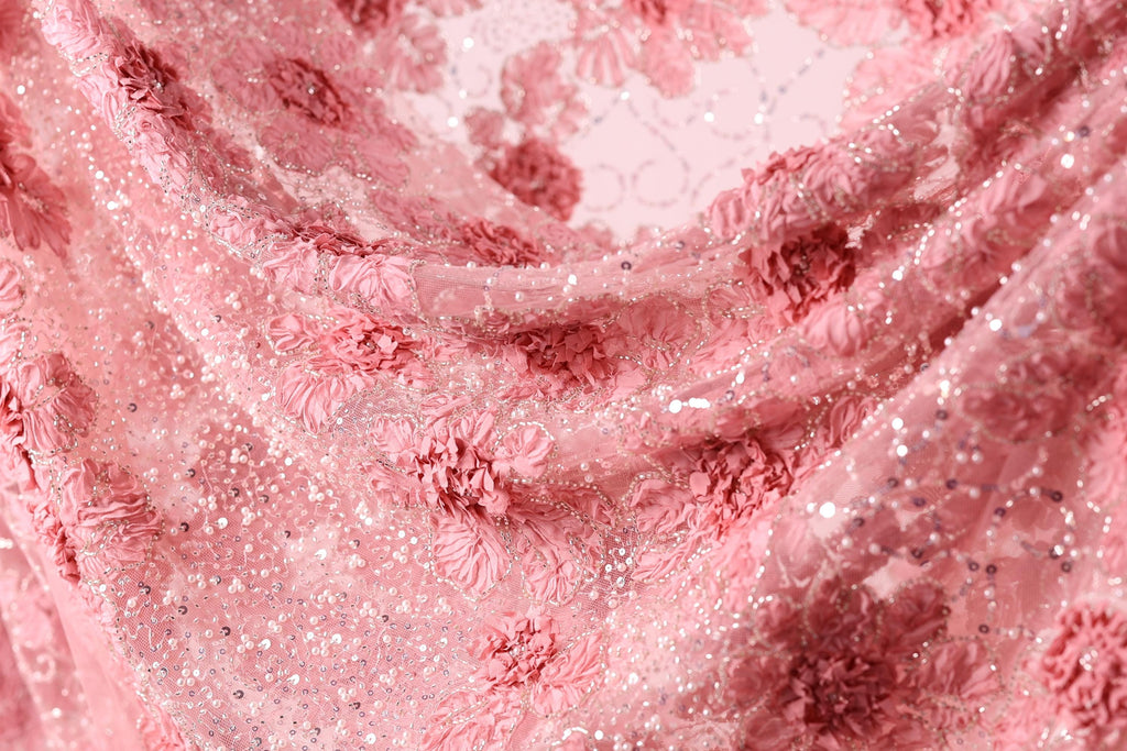 The image is a close-up of a delicate, pink fabric with a floral pattern. The fabric appears to be made of lace or a similar delicate material, with small flowers or petals visible throughout the fabric. The image does not contain any text or other discernible objects, and the focus is solely on the intricate details of the fabric.