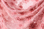 The image is a close-up of a delicate, pink fabric with a floral pattern. The fabric appears to be made of lace or a similar delicate material, with small flowers or petals visible throughout the fabric. The image does not contain any text or other discernible objects, and the focus is solely on the intricate details of the fabric.
