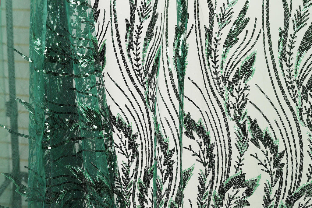 a close-up view of a fabric with a repeating pattern of green and black leaf-like designs. The pattern is intricate and detailed, with the leaves appearing to be made of sequins or beads. The fabric is predominantly white, providing a contrast to the green and black leaf motif. The image does not contain any text or other discernible objects, and the focus is solely on the leaf-like pattern on the fabric.