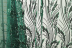a close-up view of a fabric with a repeating pattern of green and black leaf-like designs. The pattern is intricate and detailed, with the leaves appearing to be made of sequins or beads. The fabric is predominantly white, providing a contrast to the green and black leaf motif. The image does not contain any text or other discernible objects, and the focus is solely on the leaf-like pattern on the fabric.