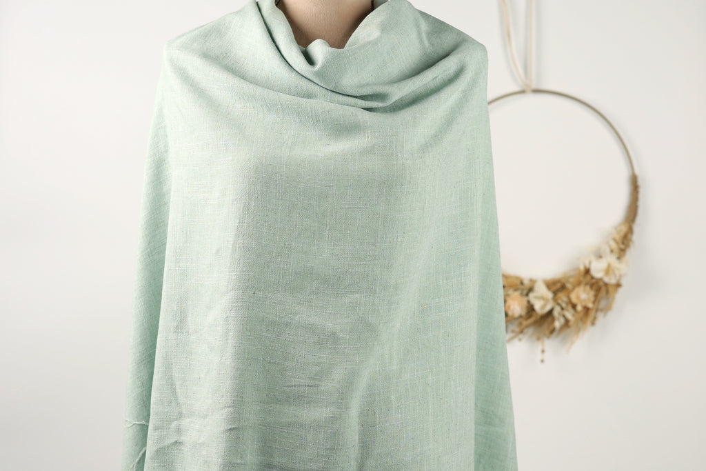 A mannequin is draped in a light green fabric, with a wreath of dried flowers hanging on the wall behind it.
