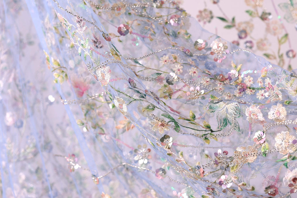 The image is a close-up of a delicate, intricately embroidered fabric with a floral pattern. The fabric appears to be made of sheer material, giving it a soft and delicate appearance. The embroidery features a variety of flowers and leaves, creating a visually appealing and intricate design. The colors used in the embroidery are predominantly pink, white, and green, which contribute to the overall beauty of the fabric.